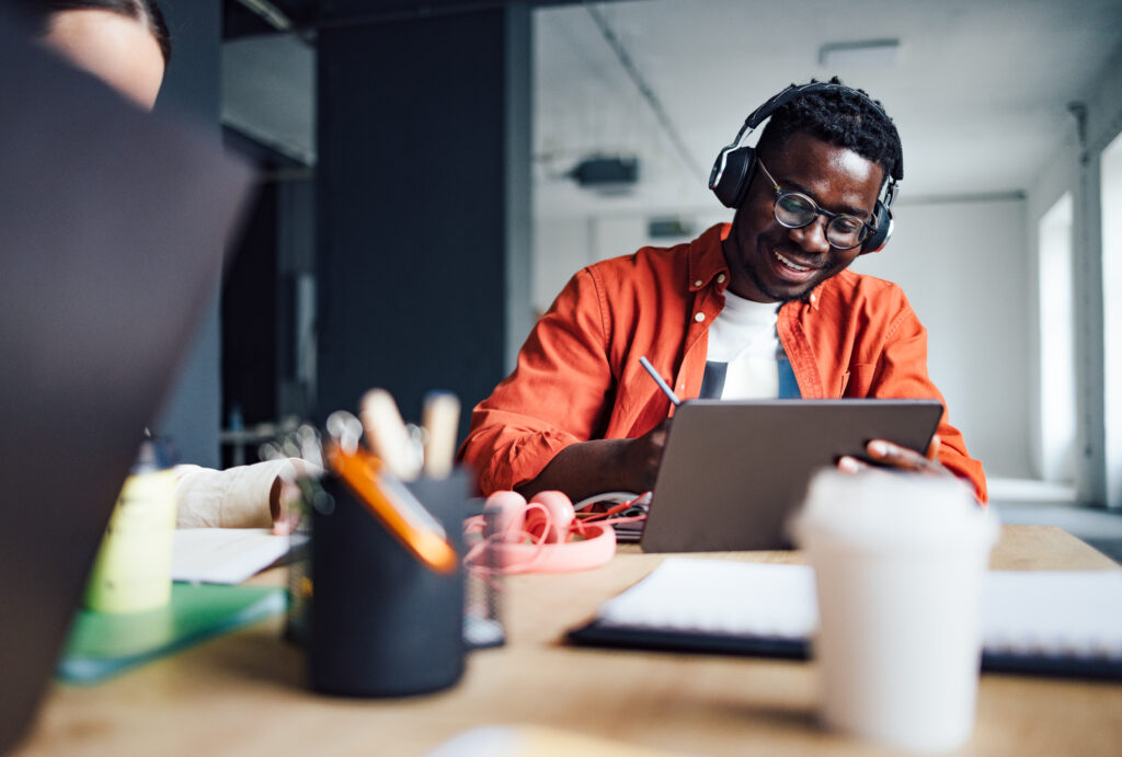 A happy looking young man is sat at a desk working on a tablet while wearing headphones.